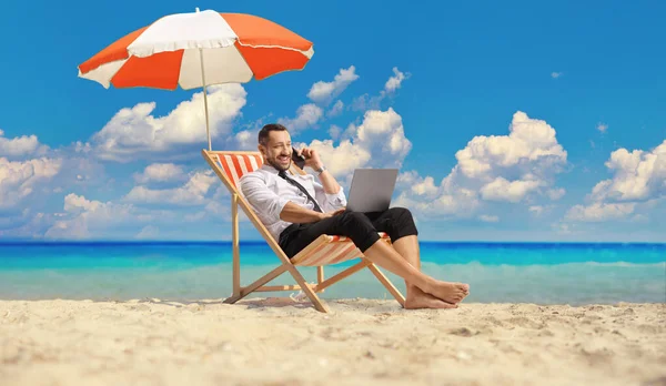 ビジネスマンは 傘の下でデッキチェアに座って ビーチでラップトップコンピュータとスマートフォンを使用しています — ストック写真