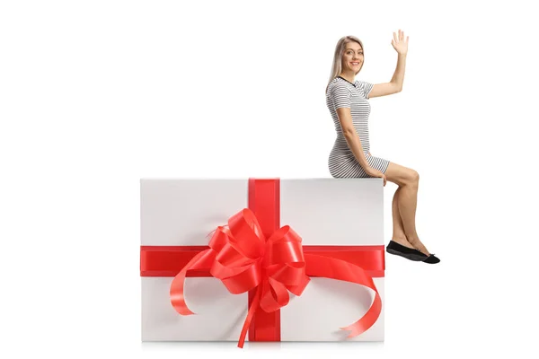 年轻女子坐在一个大礼品盒上 在白色的背景上孤零零地挥动着手 — 图库照片