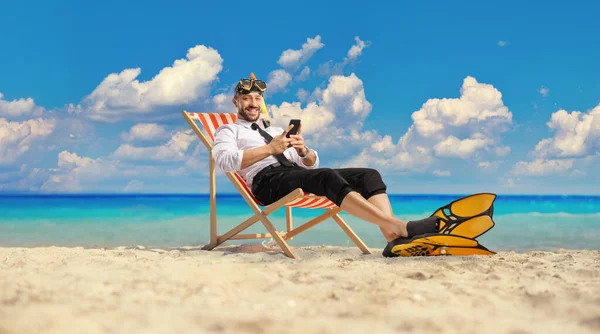 一个带着潜水鳍和面具的商人坐在海边的摇椅上 用智能手机 — 图库照片