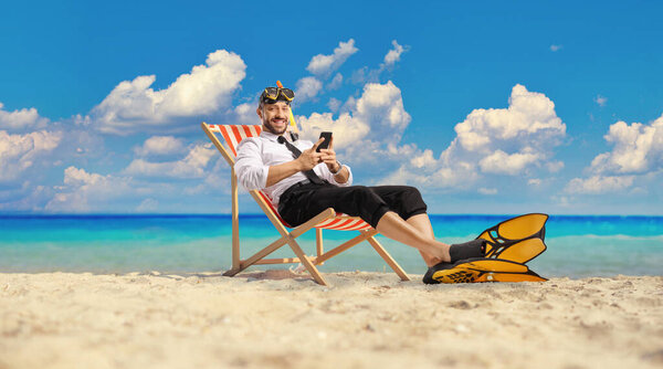 Businessman with snorkelling fins and mask sitting on a bech chair by the sea and using a smartphone