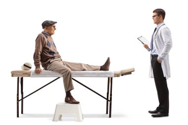 Elderly man with an injured leg sitting at a physical therapy bed and talking to a doctor isolated on white background
