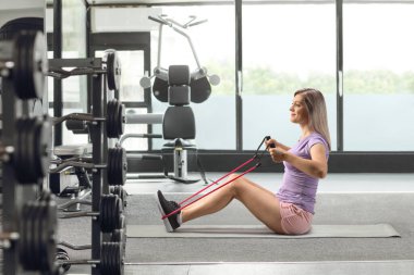 Young female sitting on a mat and exercising with a resistance band at a gym