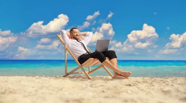 Tişörtlü ve pantolonlu bir adam sahilde dinleniyor ve dizüstü bilgisayara bakıyor.