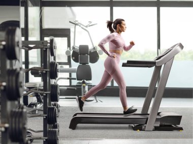 Spor salonunda koşu bandında koşan bir kadının tam profil fotoğrafı.