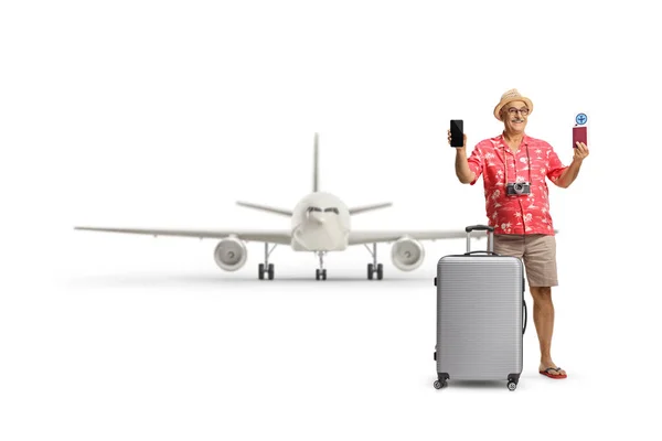 白い背景に隔離された飛行機の前にパスポートとスマートフォンを示すスーツケースを持つ成熟した男性の観光客 — ストック写真