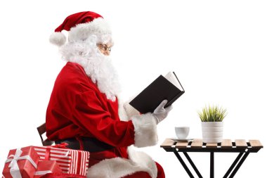 Noel Baba kahve masasında oturup beyaz arka planda izole edilmiş bir kitap okuyor.
