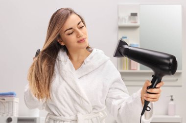 Bornozlu bir kız banyoda saç kurutma makinesiyle saçını temizliyor.