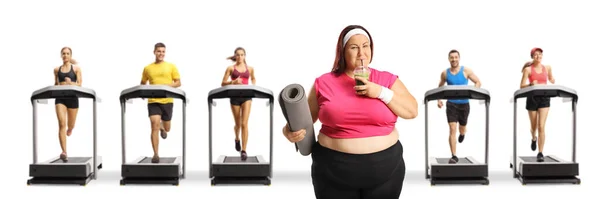 体重超标的女人喝着健康的绿色冰沙 在被白色背景隔离的跑步机上跑步的人面前拿着一块锻炼垫 — 图库照片