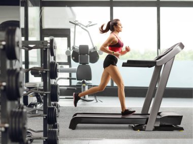 Spor salonundaki koşu bandında egzersiz yapan genç bir kadının tam boy profil fotoğrafı.