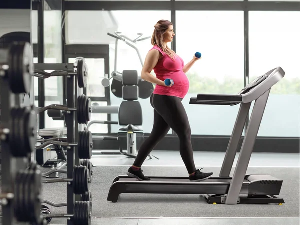 Full length profile shot of a pregnant woman walking on a treadmill with dumbbells in her hands at a gym
