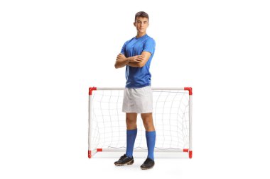 Mavi formalı genç bir futbolcunun tam boy portresi beyaz arka planda izole edilmiş mini bir kalenin önünde kollarını kavuşturmuş poz veriyor.