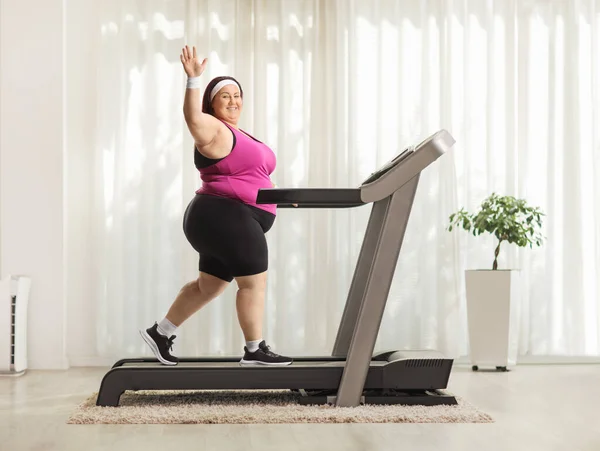 Full length profile shot of an overweight woman working out on a treadmill at home and waving at camera