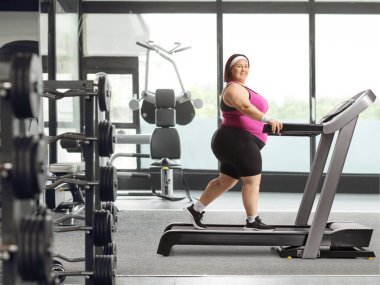 Gülümseyen şişman kadın spor salonunda koşu bandında yürüyor.