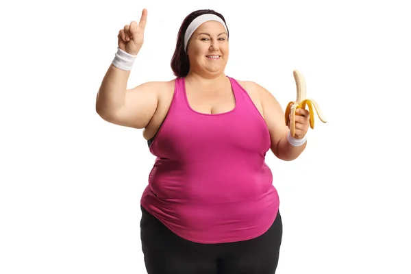 Веселая Толстая Женщина Спортивной Одежде Держит Банан Указывает Изолированный Белом — стоковое фото