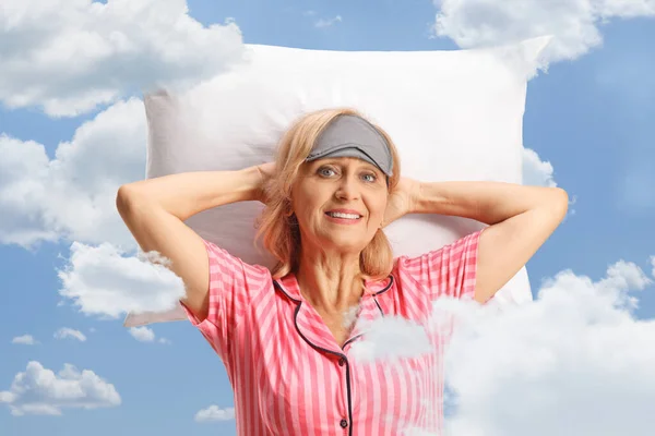 Mature woman in pajamas with a sleeping mask resting over a pillow among clouds on a blue sky