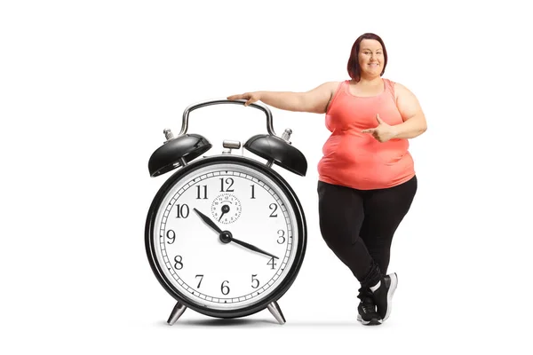 身穿运动服 面带微笑的胖女人指着一个被白色背景隔开的闹钟 — 图库照片