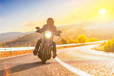 Açık bir yolda gün batımı ve motosiklet süren kasklı bir adam.