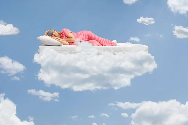 Woman in pajamas sleeping on a mattress and floating on a cloud up in the sky