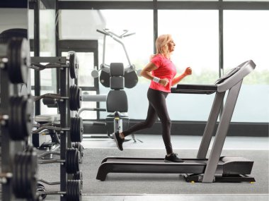 Yetişkin bir kadının spor salonunda koşu bandında koşarken tam boy profil fotoğrafı.
