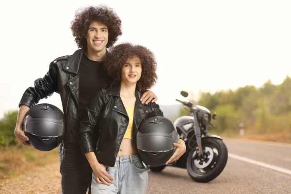 Couple in leather jackets holding helmets in front of a parked motorbike on an open road