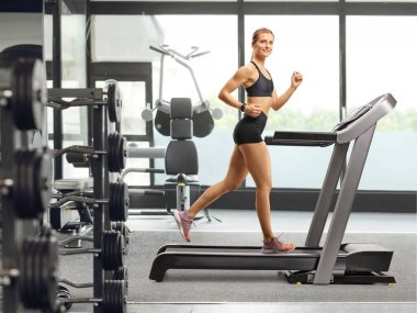 Spor salonunda koşu bandında koşan ve kameraya bakan çekici bir kadının tam boy fotoğrafı.