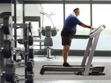 Yetişkin bir adamın spor salonundaki koşu bandındaki güç düğmesine bastığı tam boy profil fotoğrafı.