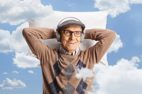Elderly man with headphones resting on a pillow and smiling among clouds