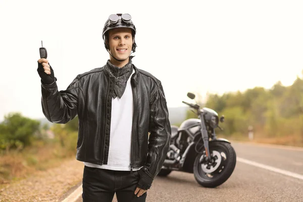 Biker in a leather jacket holding keys next to a parked chopper on the road