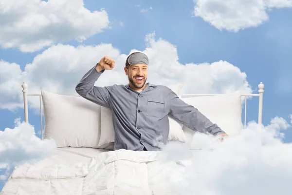 Happy young man with a sleeping mask stretching in a comfortable bed up in the clouds