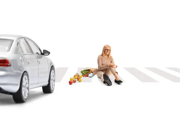 一个拿着购物袋膝盖受伤的女人坐在大街上 坐在一辆白色背景隔离的汽车前面 — 图库照片