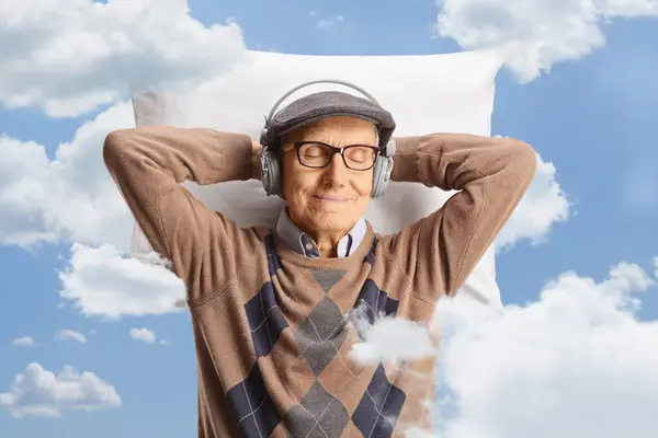 Elderly man resting on a pillow up in the clouds and listening to music with headphones