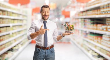 Küçük bir alışveriş arabası tutan ve bir süpermarketi işaret eden genç adam. 