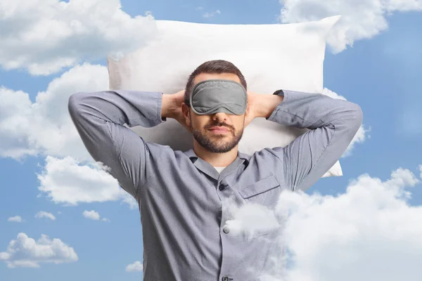 Man in pajamas with a sleeping mask laying on a pillow between clouds in the sky
