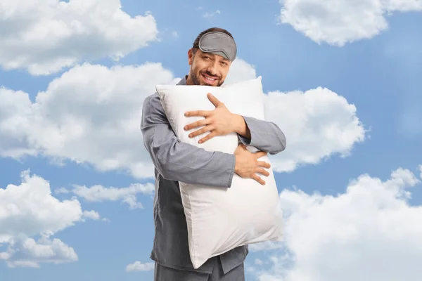 Happy man in pajamas with a sleeping mask hugging a pillow and smiling with clouds and blue sky in the background