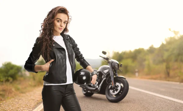 Female biker in a leather jacket holding a helmet and posing on the roa