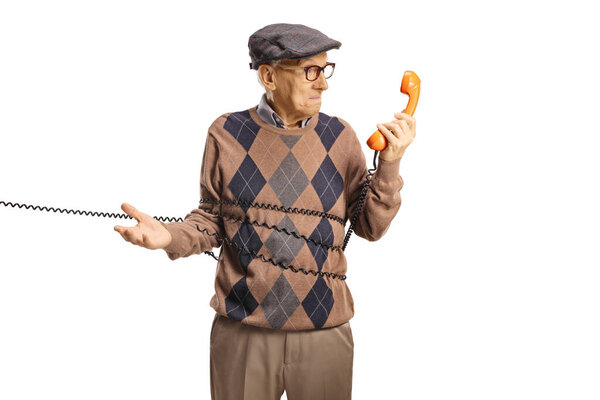 Пожилой человек запутался в кабеле от винтажного ротационного телефона, глядя на телефон, изолированный на белом фоне