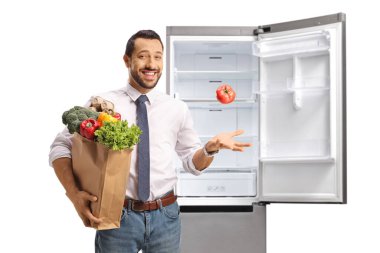 Bakkal çantası tutan neşeli bir genç adam ve beyaz arka planda izole edilmiş bir buzdolabının önünde domatesle hokkabazlık yapıyor.