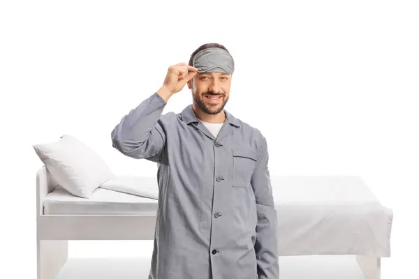 白い背景に隔離されたベッドの前に立っている睡眠マスクとパジャマの陽気な男 — ストック写真