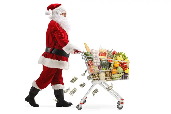 サンタクロースのウォーキングとショッピングカートを押す完全な長さのプロフィールショット 白い背景で孤立した食べ物とお金 — ストック写真
