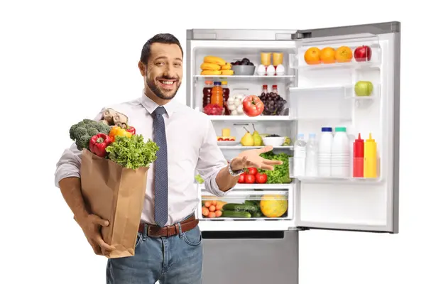 年轻快乐的职业男子拿着购物袋站在一个开着的冰箱前 冰箱被白色的背景隔开 — 图库照片