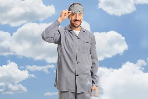 Young man in pajamas with a sleeping mask standing between clouds in the sky