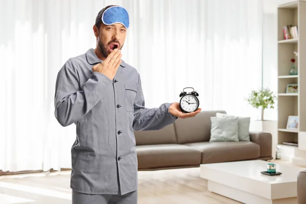 Young man in pajamas with a sleeping mask holding an alarm clock and yawning at home in a living room