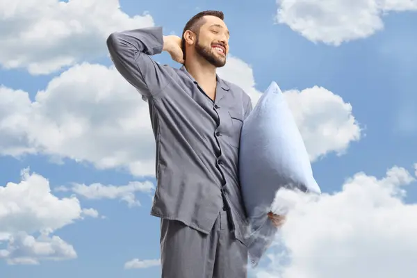 Young man in pajamas holding a pillow and stretching with blue sky and clouds in the background