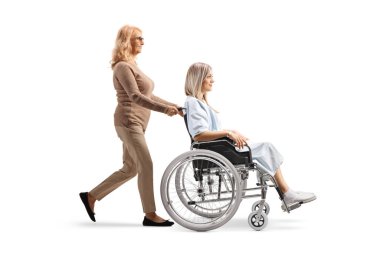 Olgun bir kadının beyaz arka planda yalnız bırakılmış tekerlekli sandalyedeki genç bir kadını iterken tam boy profil fotoğrafı.