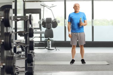 Spor kıyafetli olgun bir adamın spor salonunda elinde plastik bir şişeyle tam boy portresi.