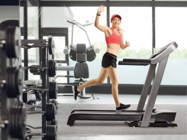 Spor salonunda koşu bandında koşan ve kameraya el sallayan bir kadın.