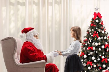 Noel Baba koltukta oturmuş ve süslenmiş bir Noel ağacıyla evde bir kızla el ele tutuşuyor.