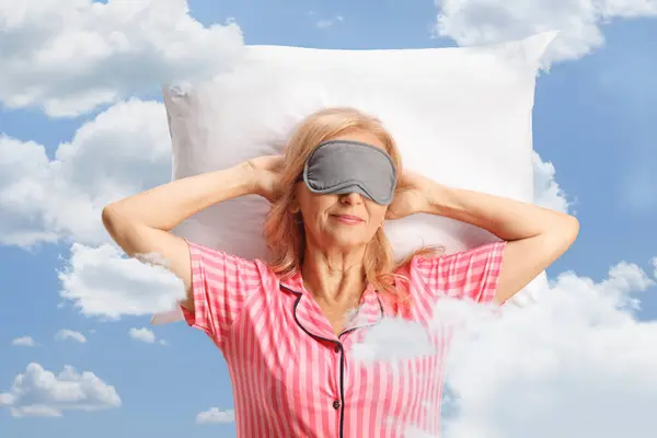 Woman in pajamas with a mask on eyes sleeping on a pillow between clouds and blue sk