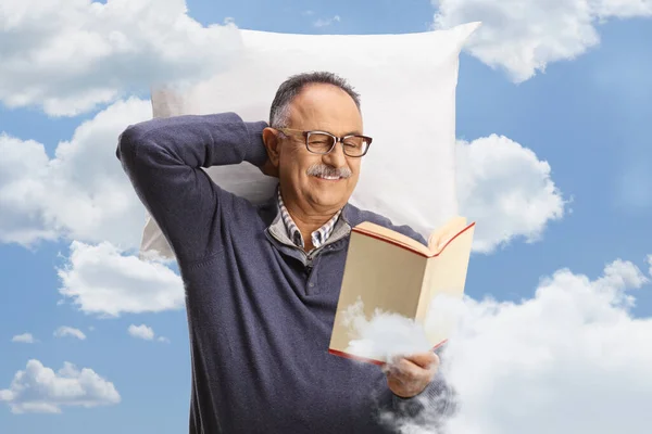 Mature man resting on a pillow and reading a book, clouds and sky in the background