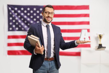 Gözlüklü genç adam kitapları tutuyor ve Amerikan bayrağının önünde A harfini taşıyor.